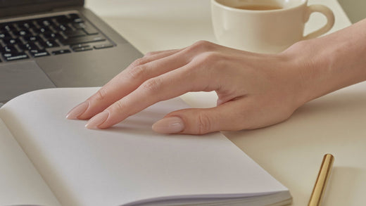 Mao com unhas press-on curtas em tom nude pousada num caderno ao lado de um teclado de portatil
