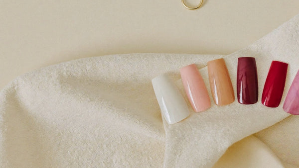 Sete unhas press-on em tons nude, rosa, coral, vinho e vermelho dispostas em arco sobre linho cru