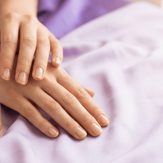 Duas mãos com unhas naturais curtas e saudáveis pousadas sobre tecido lilás