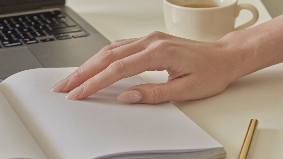 Mao com unhas press-on curtas em tom nude pousada num caderno ao lado de um teclado de portatil
