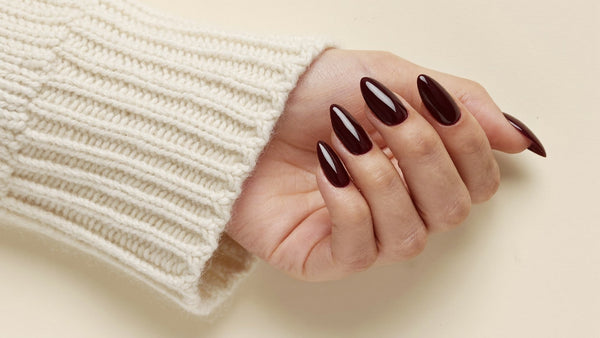 Grande plano de unhas press-on amendoadas em vinho profundo a sair de uma camisola de la creme