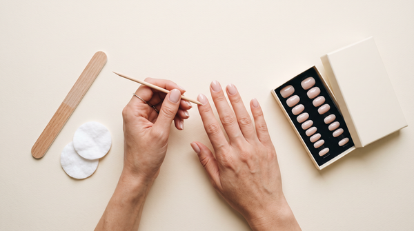 Manicure em casa passo a passo: mãos a empurrar a cutícula com pau de laranjeira, lima, discos de algodão e caixa de press-on nude